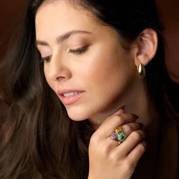 Woman wearing 14K yellow earrings and a colorful ring, with a warm and soft background.