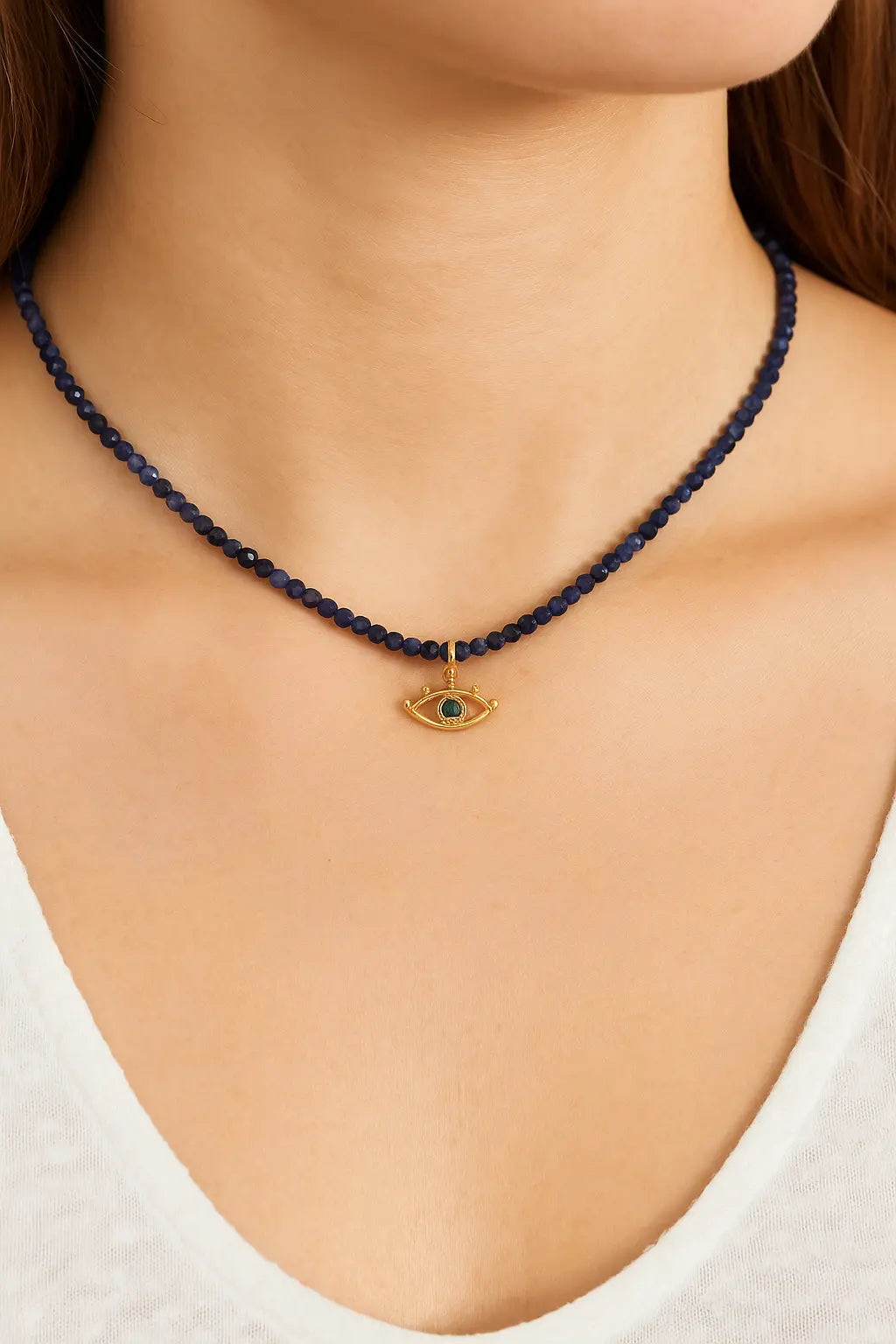 Sapphire beaded necklace with a gold and green evil eye pendant on a person wearing a white top.